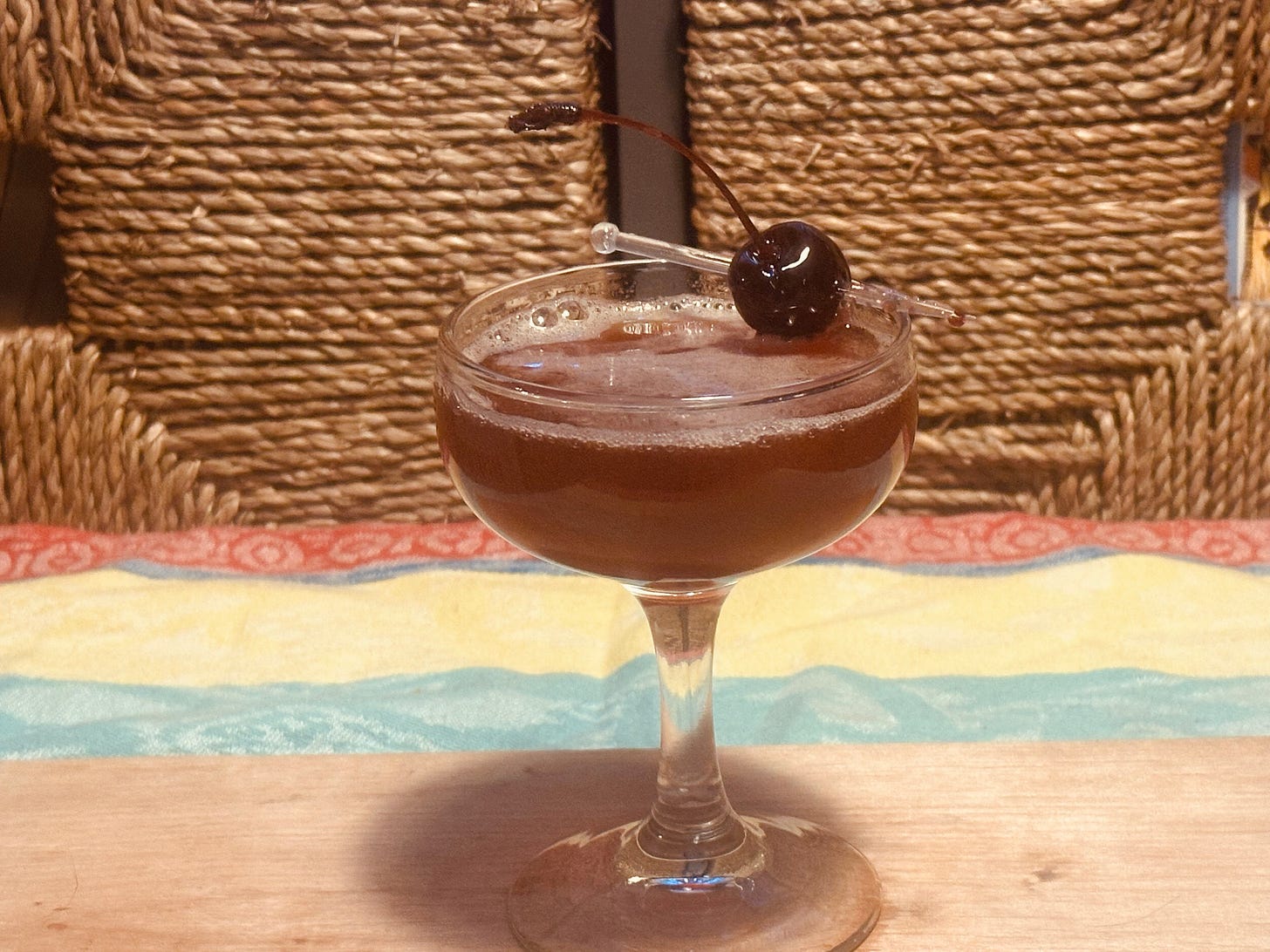 A stemmed cocktail glass with a brownish red drink in it, garnished with a succulent cherry, photographed on a wood plank atop a pastel yellow and green tablecloth. 