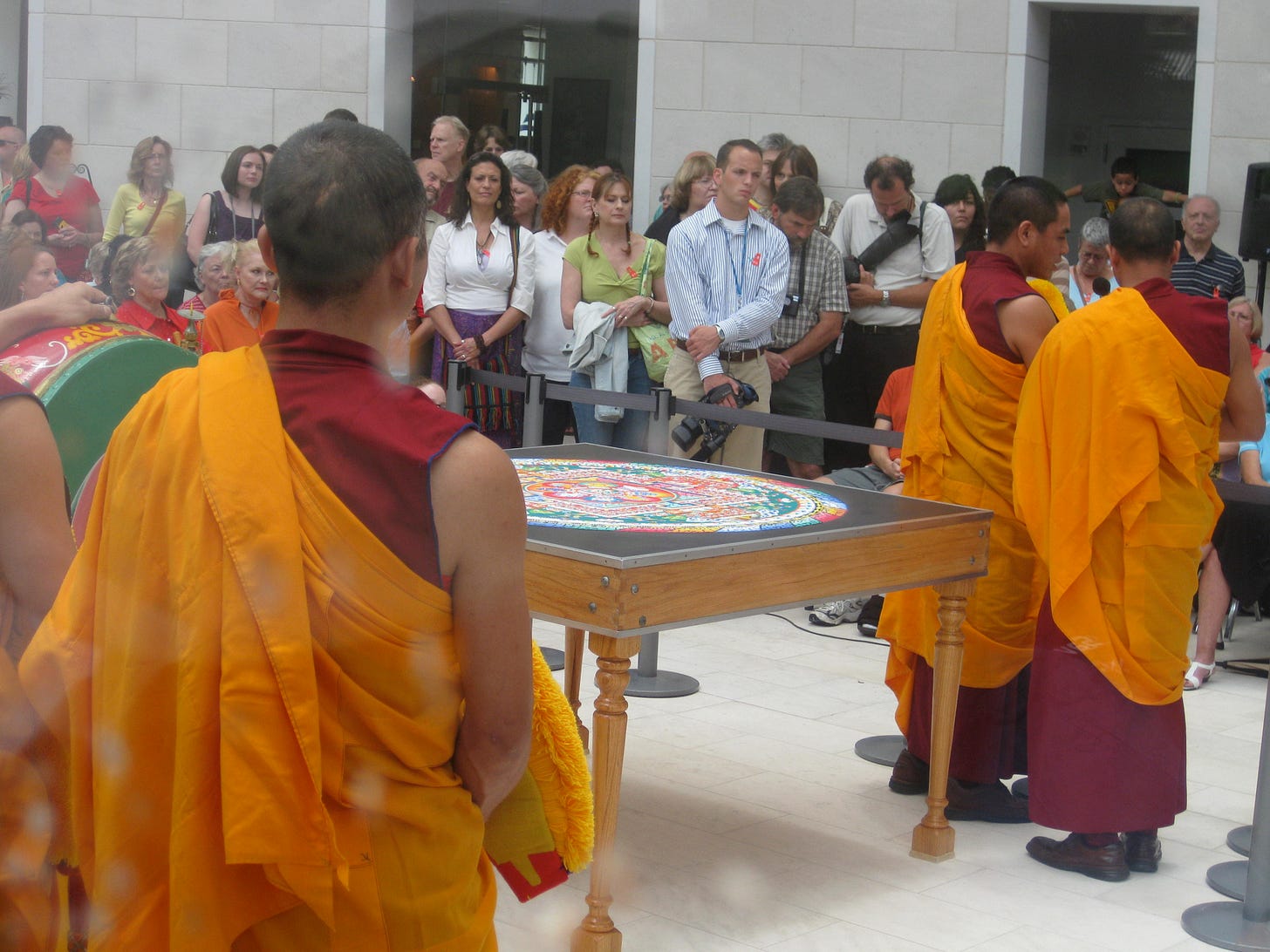 The backs of three monks, their torsos wrapped in marigold colored robes, facing a large table with a sand mandala, with a crowd of people watching beyound a guard-rope.