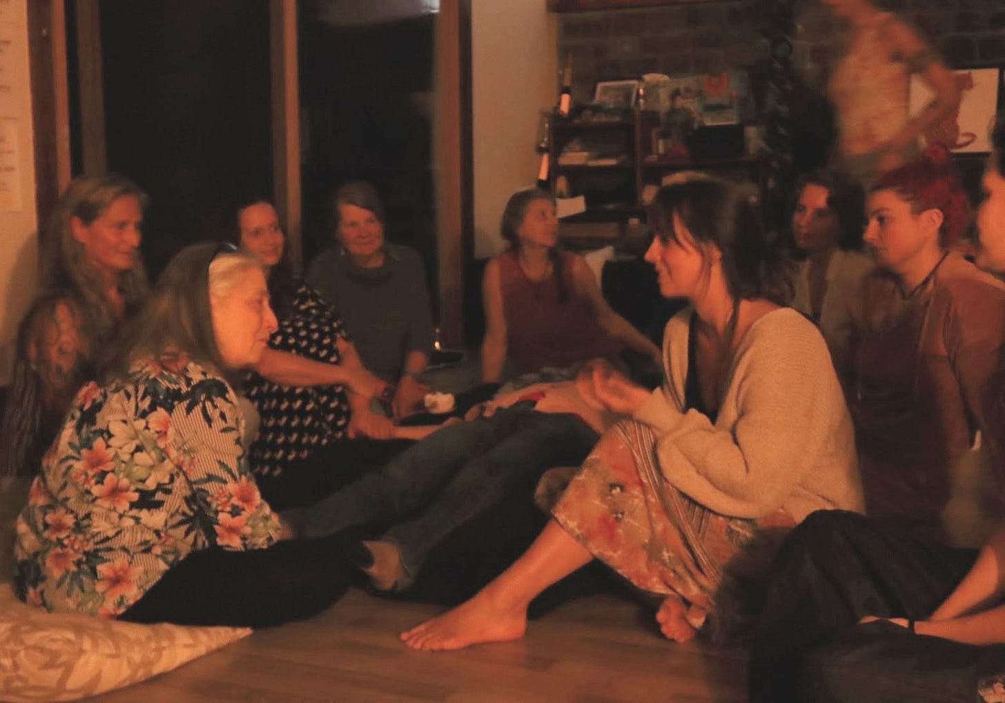 An indoor evening circle of women of several generations sitting on the floor in conversation, soft low light