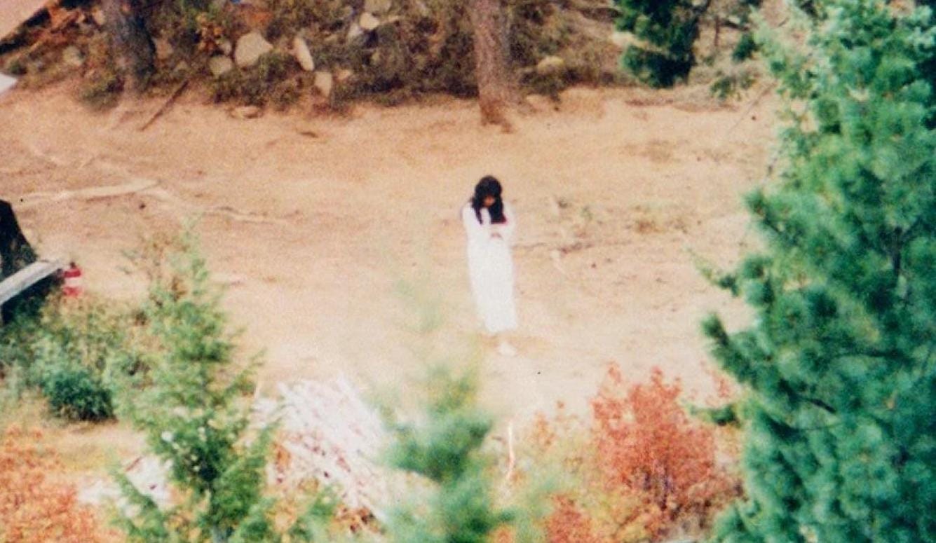 A photograph of a young woman in a forest clearing. She wears a white dress and sneakers and has long, dark hair. A photograph of a young woman in a forest clearing. She wears a white dress and sneakers and has long, dark hair.