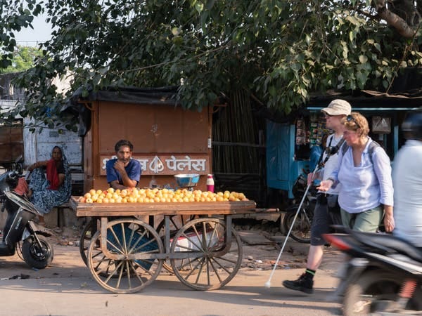 A woman walks next to a man who is holding a white cane out in front of him, while the tow of them pass a street vendor selling orange fruit displayed on a wooden cart. Lush greenery is visible in the background.