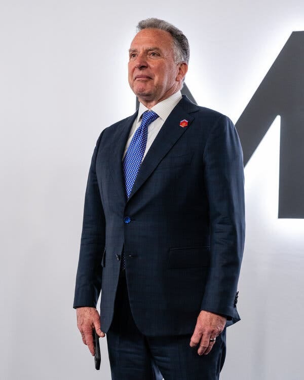 Steve Witkoff standing in front of a white background wearing a dark suit and blue tie and holding a smartphone.
