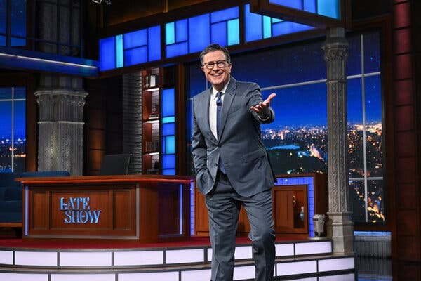 A smiling man in a blue suit and tie over a white shirt gestures down at the camera. Behind him is a desk with a log that reads “Late Show.”