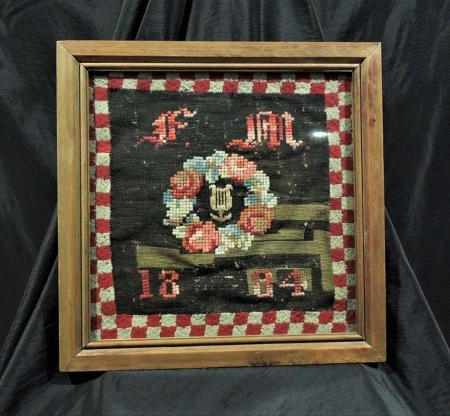May include: A framed sampler with a black background and a red and white checkered border. The sampler features a floral wreath with a lyre in the center and the text "F.M. 18-84".