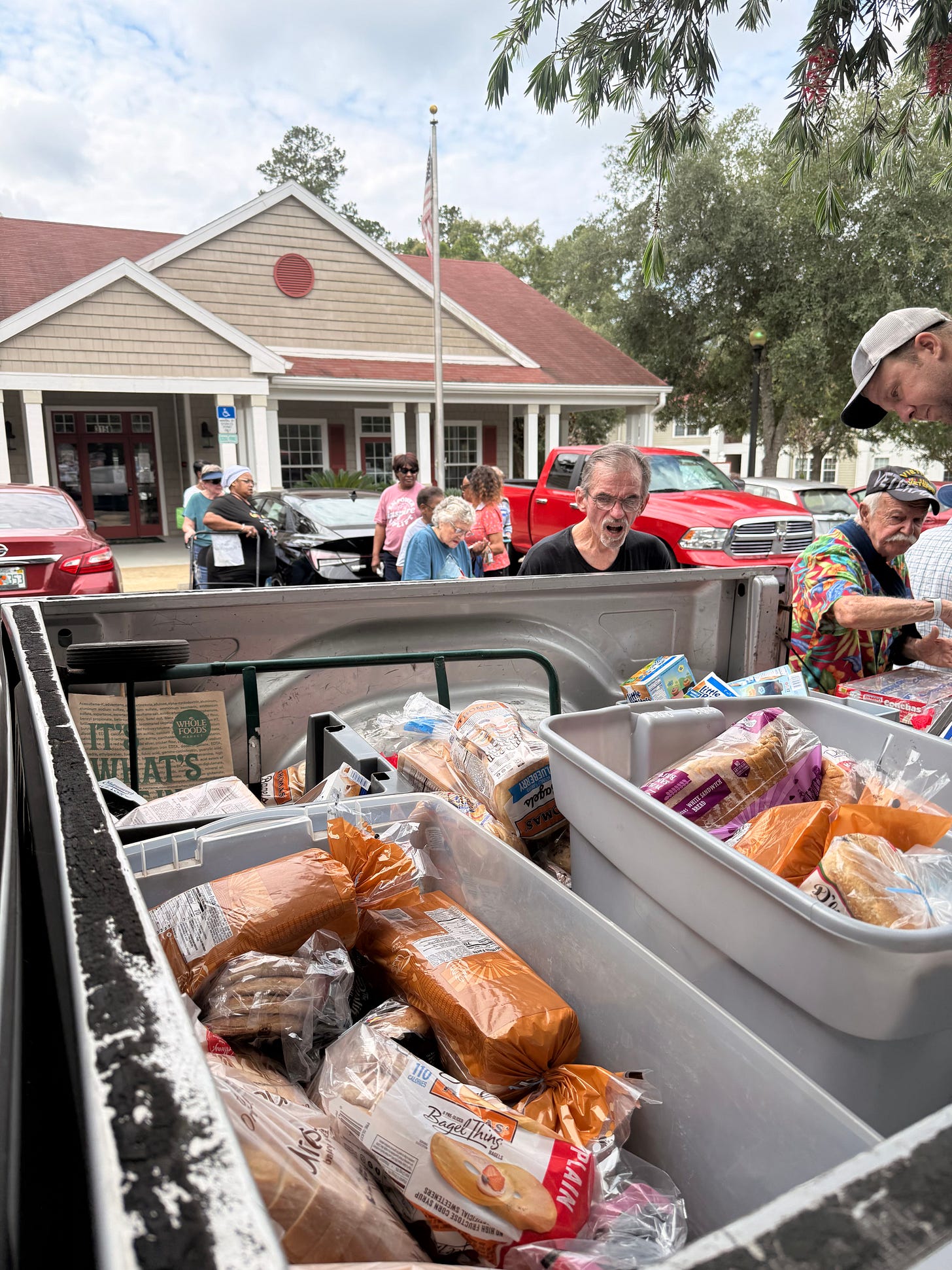 A pickup truck bed filled with packaged bread and groceries sits in the foreground, including a Whole Foods Market bag and Thomas’ Bagel Thins, while several people stand and sort items in a parking lot outside a community-style building with an American flag on a pole; cars and a red pickup truck are parked nearby under trees.