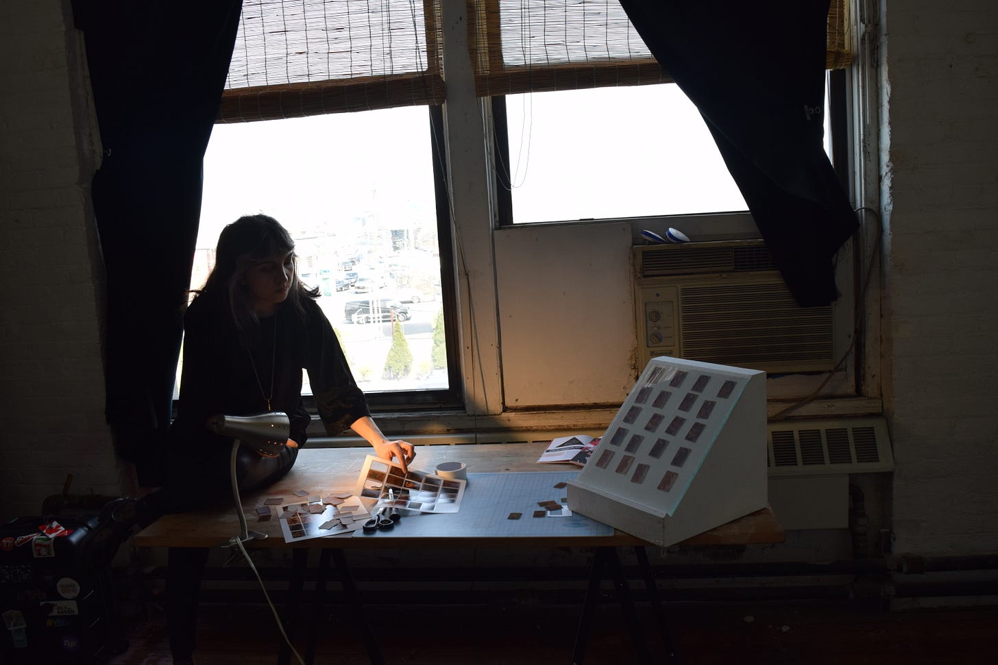 A person in black blouse is arranging prints on a studio desk in front of windows..