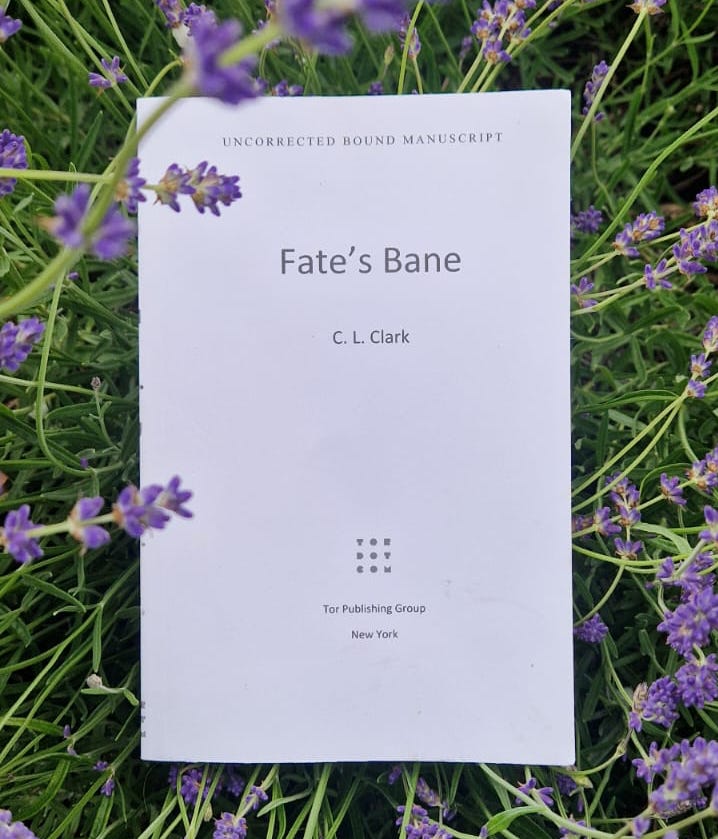 a picture of an advanced bound manuscript of Fate's Bane by C. L. Clark, lying in green grass with purple flowers--lavender??
