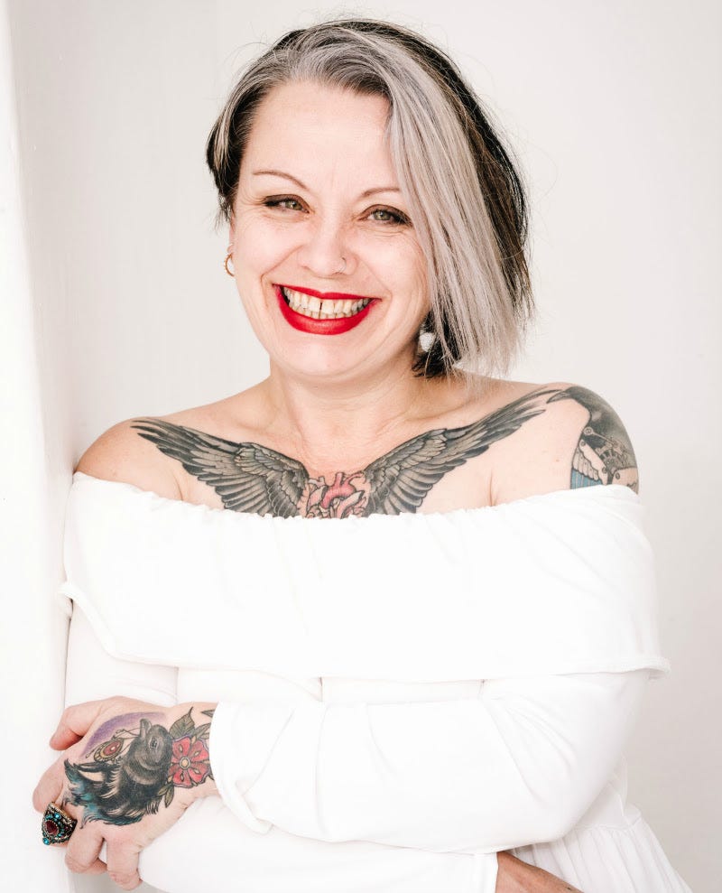 Violet's wearing red lipstick and a white off the shoulder top. She has tattoos, a white stripe in the front of her hair and a big smile. 