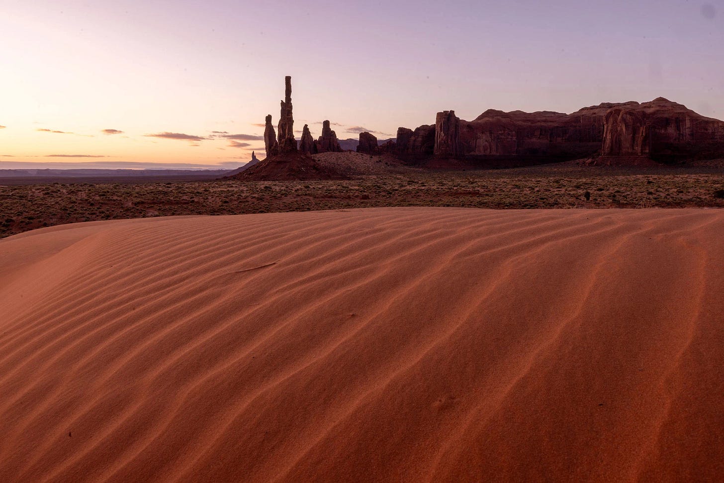 Pre-dawn light over rippled dunes at Sand Springs in Monument Valley, with the Totem Pole and sandstone monoliths in the distance where Ancestral Puebloans once lived.
