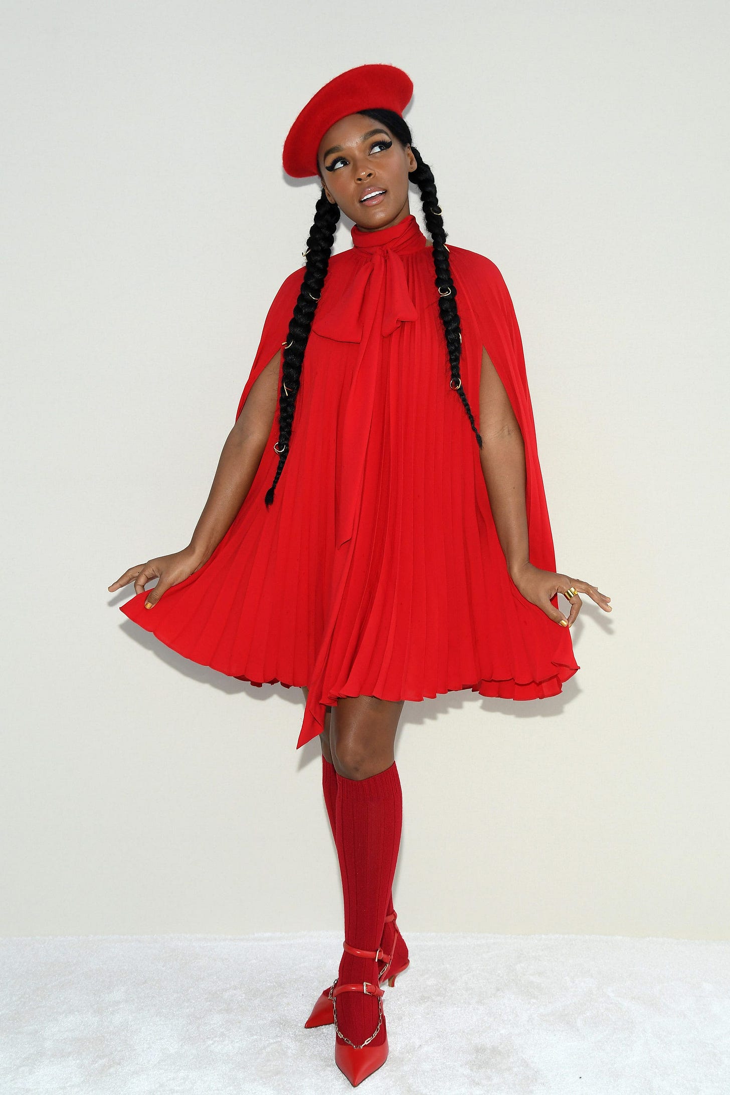 Janelle Monae poses for the camera, gently lifting her face to the left with eyes upward in charming braided pig tails. She wears a monotone outfit in postbox red: knee high stockings, a beret, a cape with a pussybow neck tie and a darling frock adorn her.
