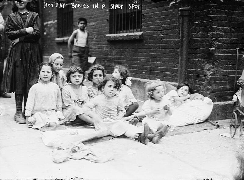 A group of children is sitting and lying down on the sidewalk in front of a brick building, seeking shade. Most are looking towards the camera. Written text at the top of the image says "Hot Day - Babies in a Shady Spot." Another child and an adult stand in the background.