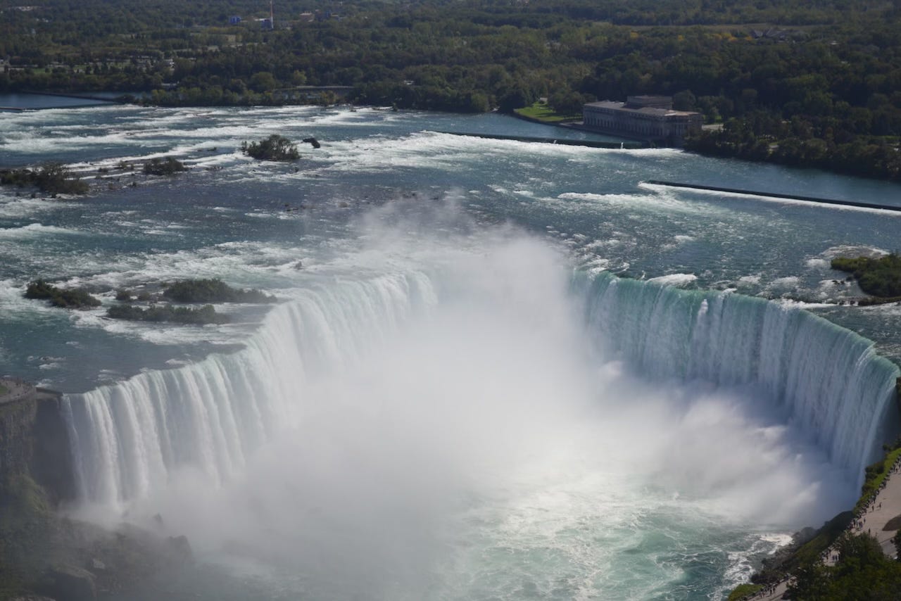 A wide aerial view of Niagara Falls, showing the massive Horseshoe Falls curving in a dramatic arc as powerful white water plunges into a mist-filled basin. The turquoise river above the falls flows around small islands before spilling over the edge, while green forest and buildings stretch across the background.