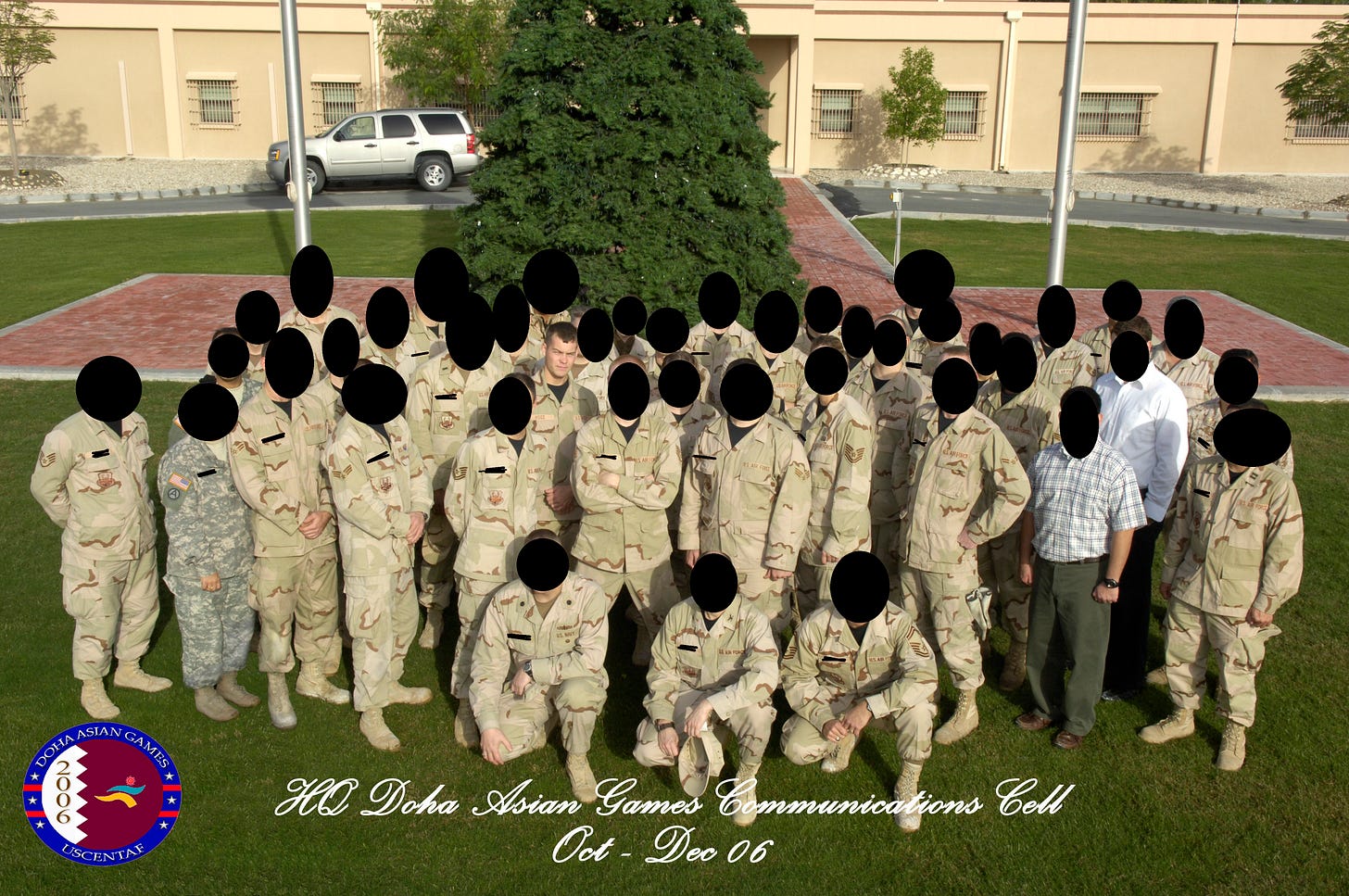 A large group photo of U.S. military personnel in desert camouflage uniforms, with a few people in different uniforms and civilian clothes, posed on a grassy lawn in front of a large evergreen tree and a beige building with barred windows; a silver SUV is parked in the background. Text at the bottom reads “HQ Doha Asian Games Communications Cell Oct - Dec 06,” and a circular “Doha Asian Games 2006” emblem appears in the lower-left corner. A large group photo of U.S. military personnel in desert camouflage uniforms, with a few people in different uniforms and civilian clothes, posed on a grassy lawn in front of a large evergreen tree and a beige building with barred windows; a silver SUV is parked in the background. Text at the bottom reads “HQ Doha Asian Games Communications Cell Oct - Dec 06,” and a circular “Doha Asian Games 2006” emblem appears in the lower-left corner.