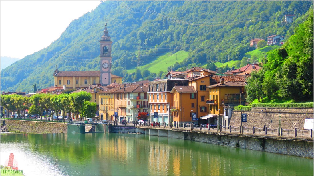 Colorful buildings and a clock tower line a tranquil river, set against a lush, green mountain backdrop. Clear sky and serene atmosphere. San Pelligrino, Italy.