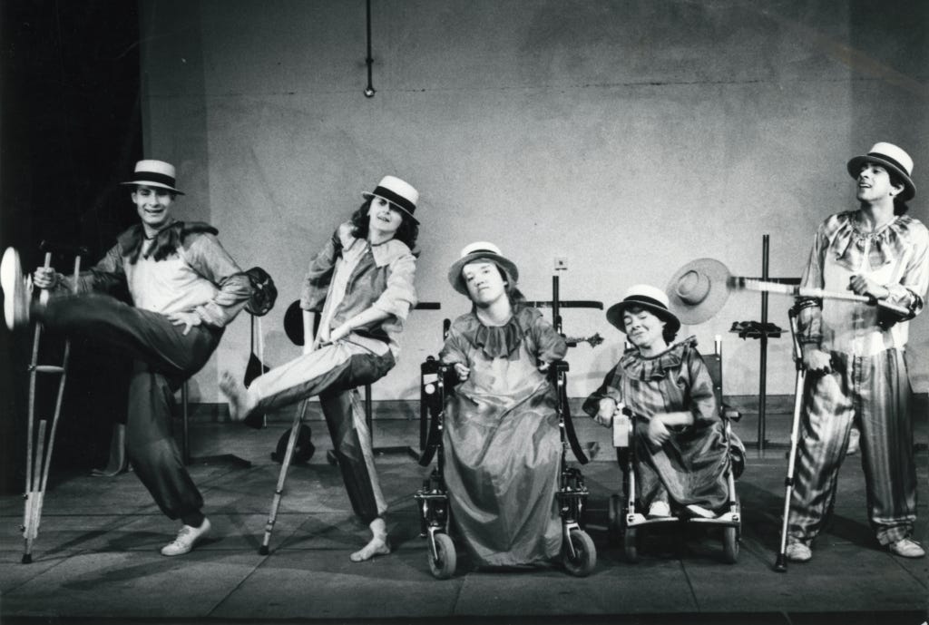 Black and white theatrical photograph of five Graeae performers posed in a chorus line formation on a stage. The performers wear costumes including boater hats and period-style clothing. From left to right: a performer seated on a raised platform with one leg extended, a standing performer leaning dynamically, a central performer in a dress posed on the floor, a performer seated in a wheelchair wearing a wide-brimmed hat, and a standing performer in patterned trousers. Black and white theatrical photograph of five Graeae performers posed in a chorus line formation on a stage. The performers wear costumes including boater hats and period-style clothing. From left to right: a performer seated on a raised platform with one leg extended, a standing performer leaning dynamically, a central performer in a dress posed on the floor, a performer seated in a wheelchair wearing a wide-brimmed hat, and a standing performer in patterned trousers.