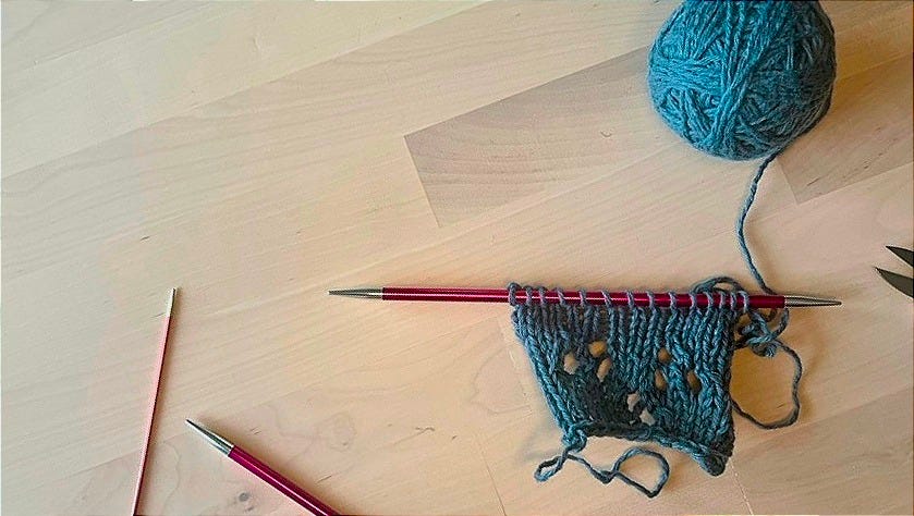 Blue-green stitches on a single straight needle on a wooden surface, showing a provisional cast on.