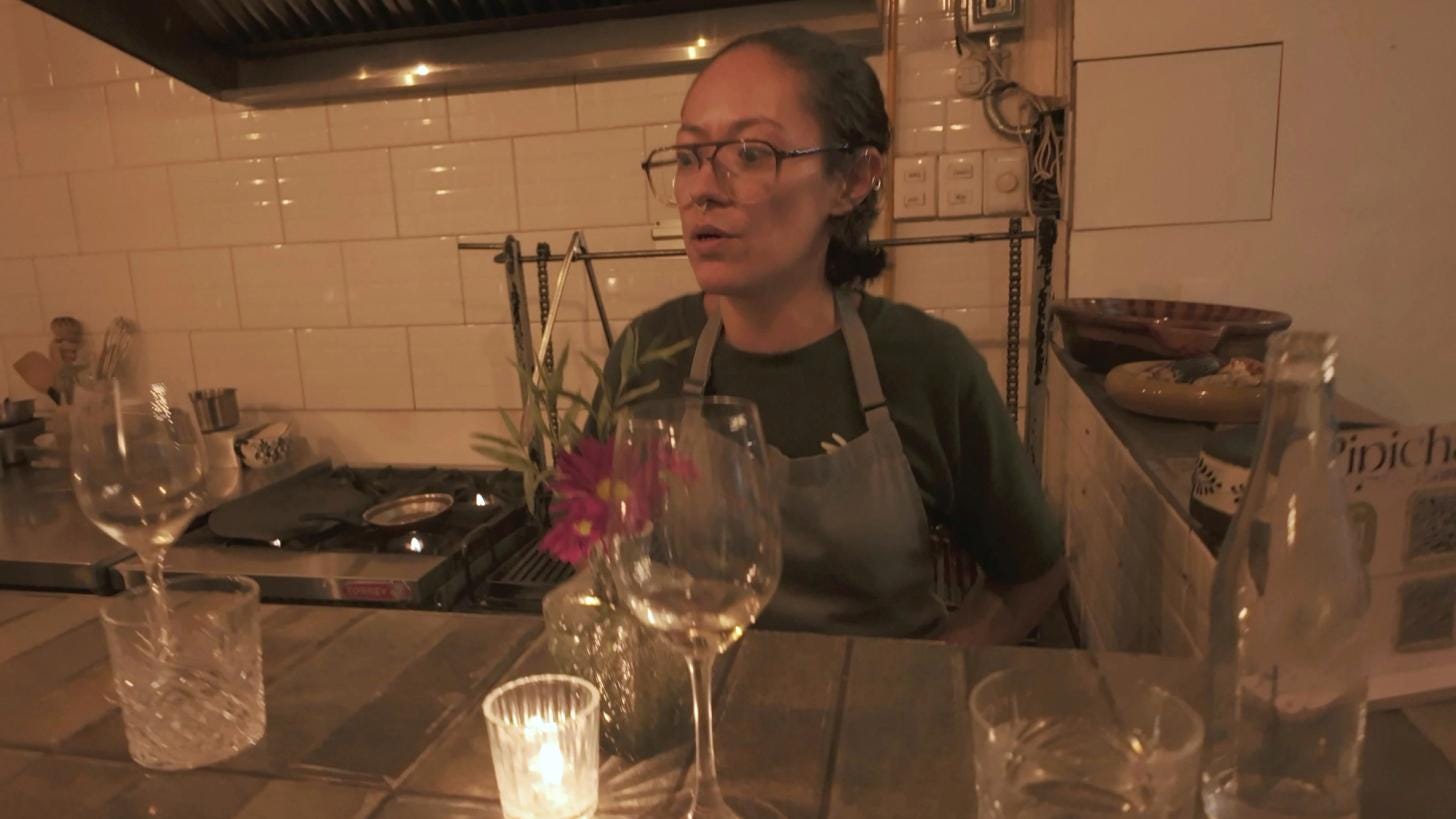 Chef Anita smiling behind the counter of the open kitchen, mid-explanation of a course. Chef Anita smiling behind the counter of the open kitchen, mid-explanation of a course.
