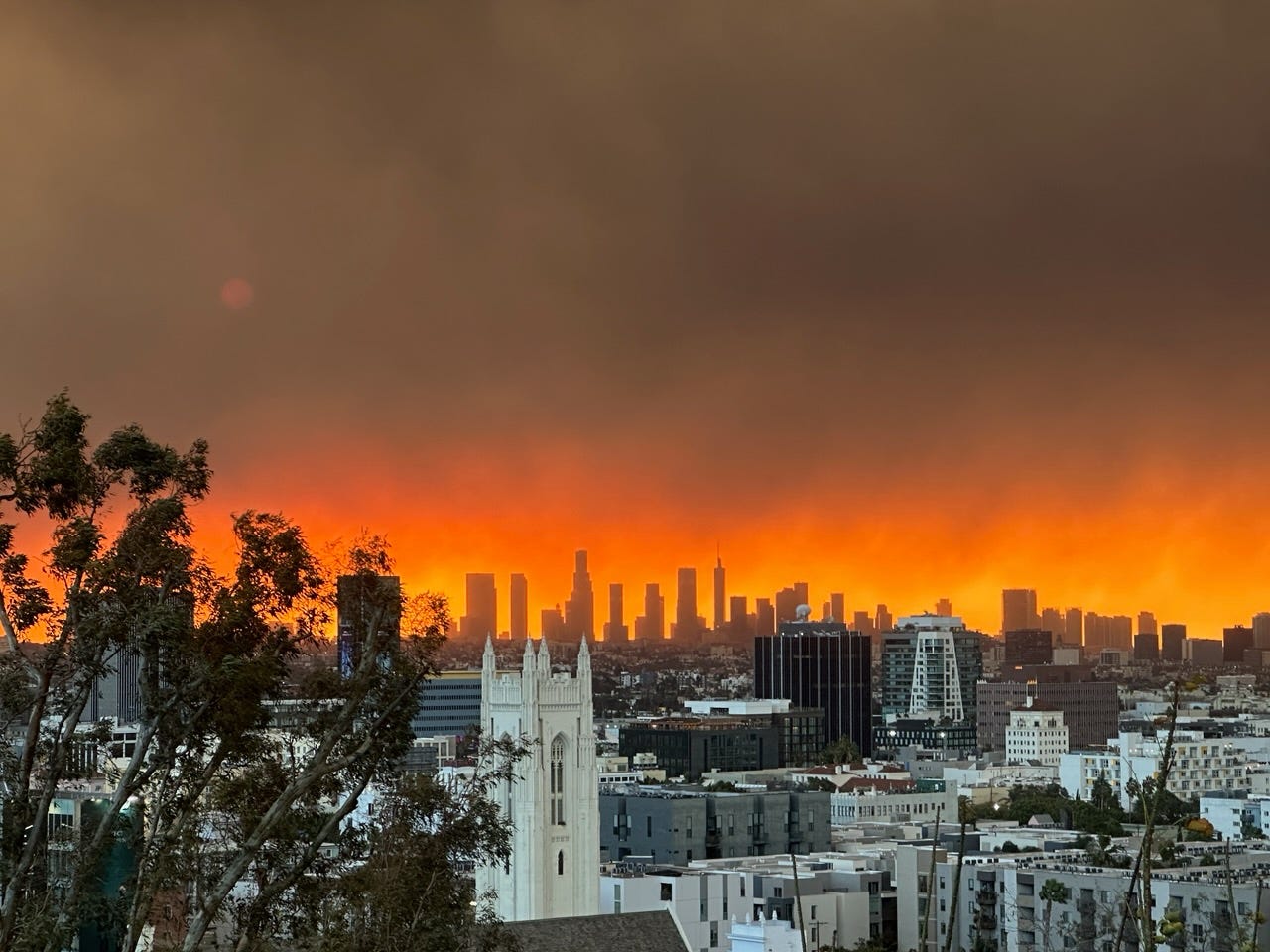 A view of Los Angeles from a rooftop during the fires. The sky behind the city skyline goes from black to red to orange, making it appear as if the entire rest of the city is in flames.