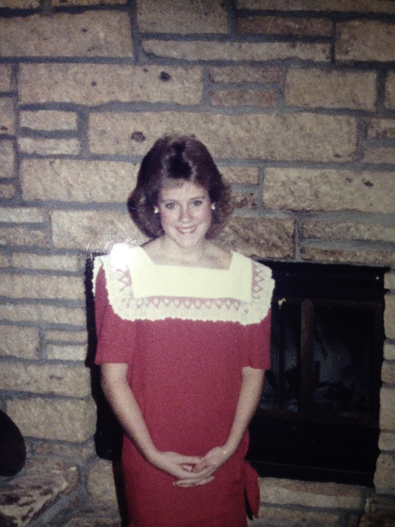 Young girl (approximately 16 years old) with dark blonde hair stands in front of a stone wall with a fireplace. She's smiling and wearing a red dress with a white pinafore collar. 