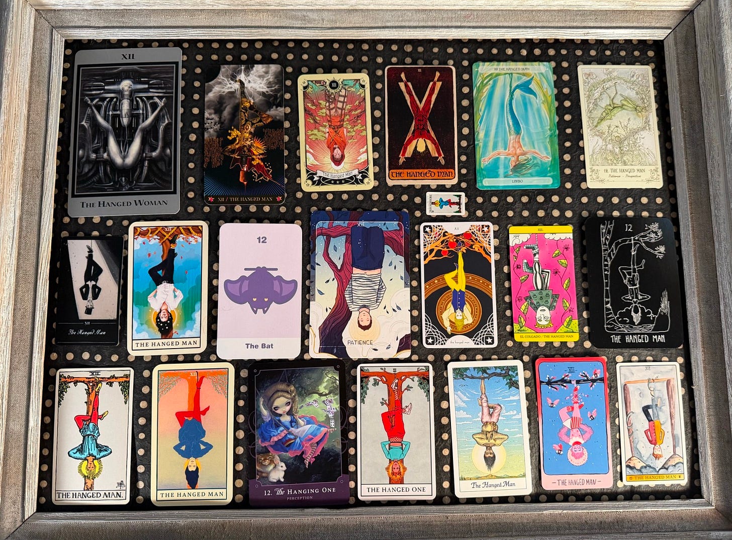 A grey wooden frame filled with about twenty tarot cards, all the same card — the Hanged Man — from different decks. Each shows a figure suspended upside down, some serene, some playful, in styles ranging from manga to Giger to pastel line art. The decks title it variously: the Hanged Man, the Hanged One, the Hanged Woman, Patience. One tiny card sits apart in its own lane.