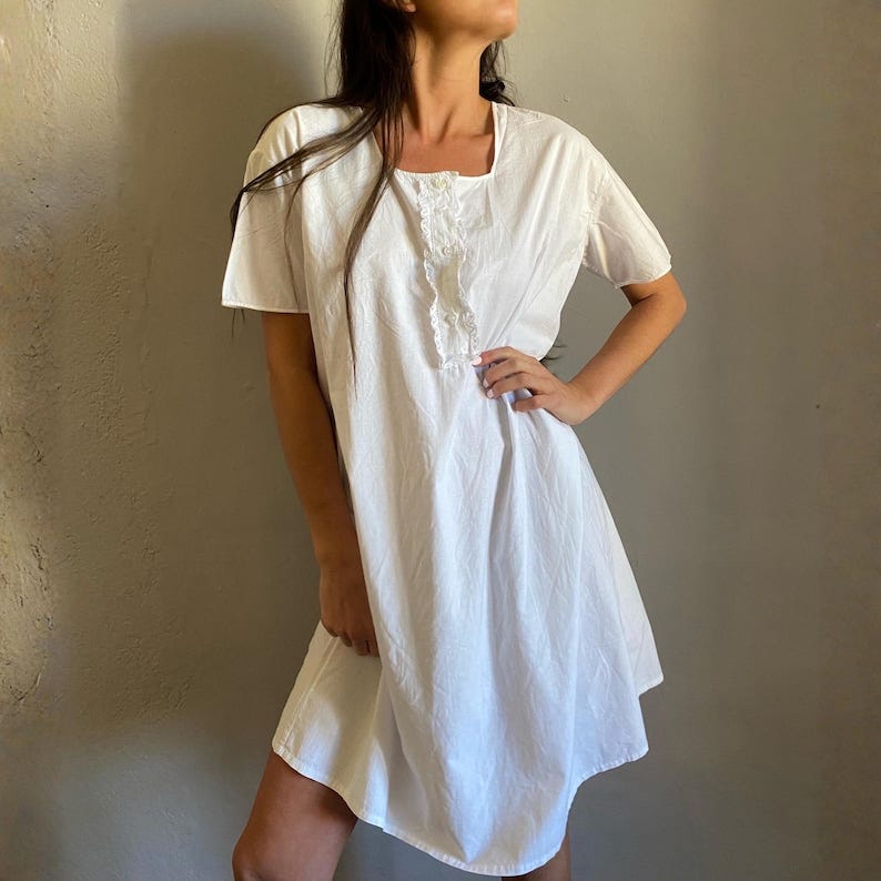 May include: A white, short-sleeved dress with a square neckline and a ruffled detail down the front. The dress is made of a lightweight fabric and has a slightly flared skirt. The model is wearing the dress.