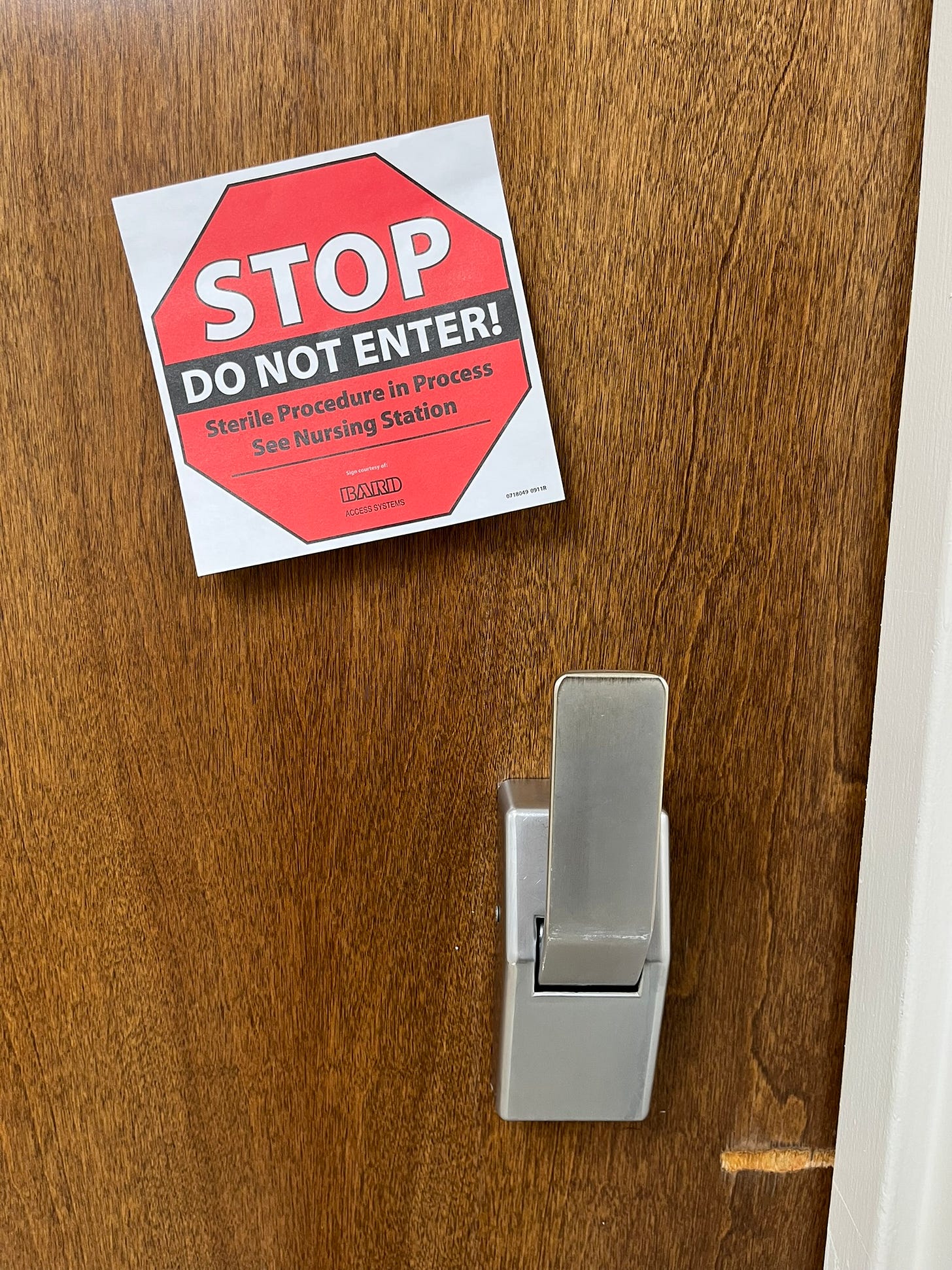 A red and white hospital sign on a wooden door reads “STOP — DO NOT ENTER! Sterile Procedure in Process. See Nursing Station.” A silver metal door handle is visible below the sign.
