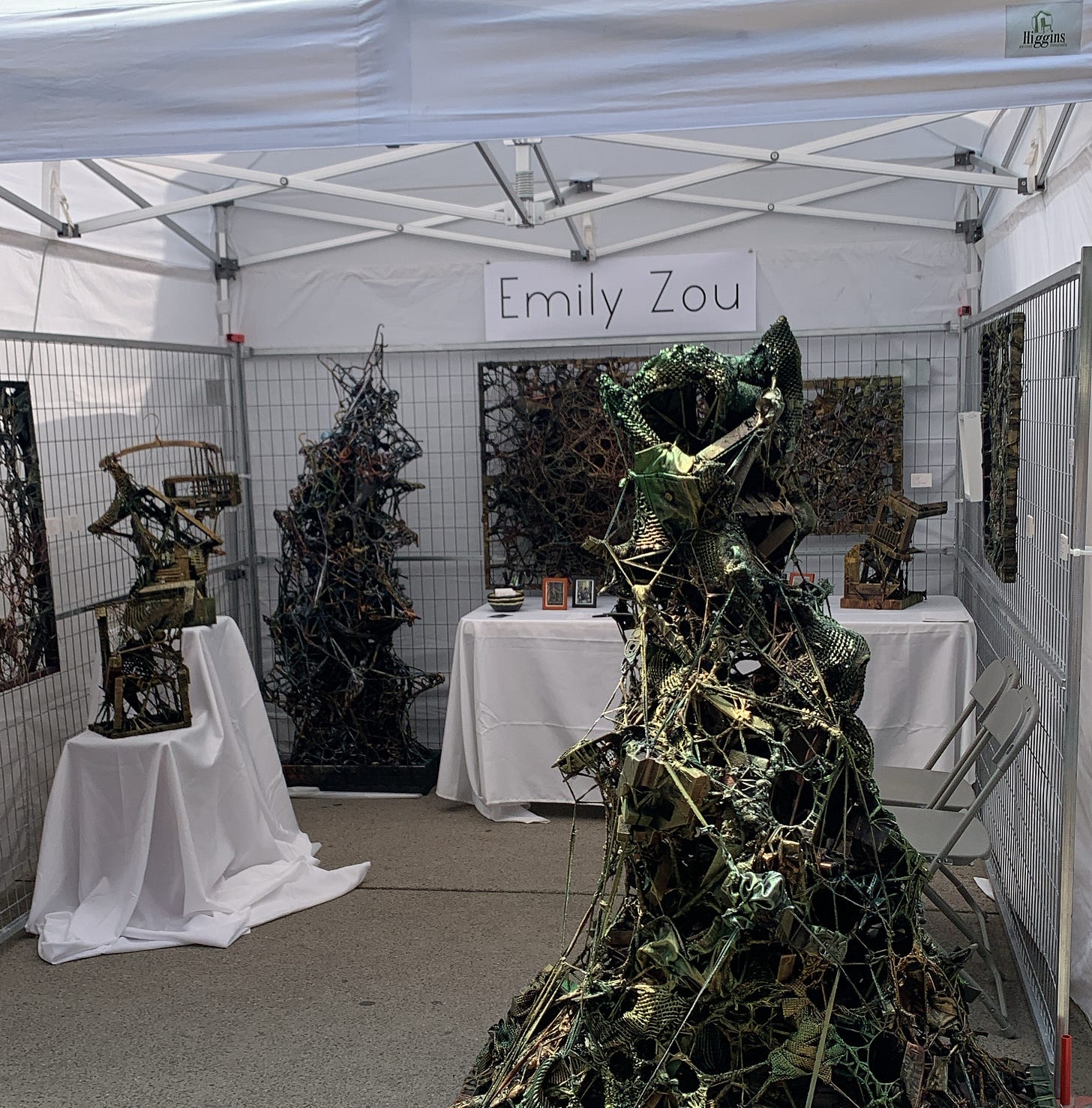 A white tent with the sign "Emily Zou," with an array of sculptures made of piles of found objects, painted dark green and gold.