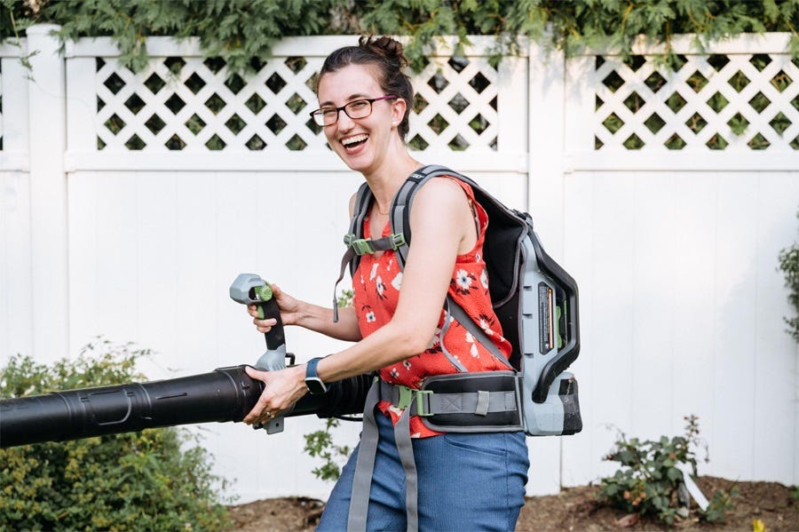 a smiling woman wearing glasses and a bright summery top tries an electric leaf blower that's strapped to her back 