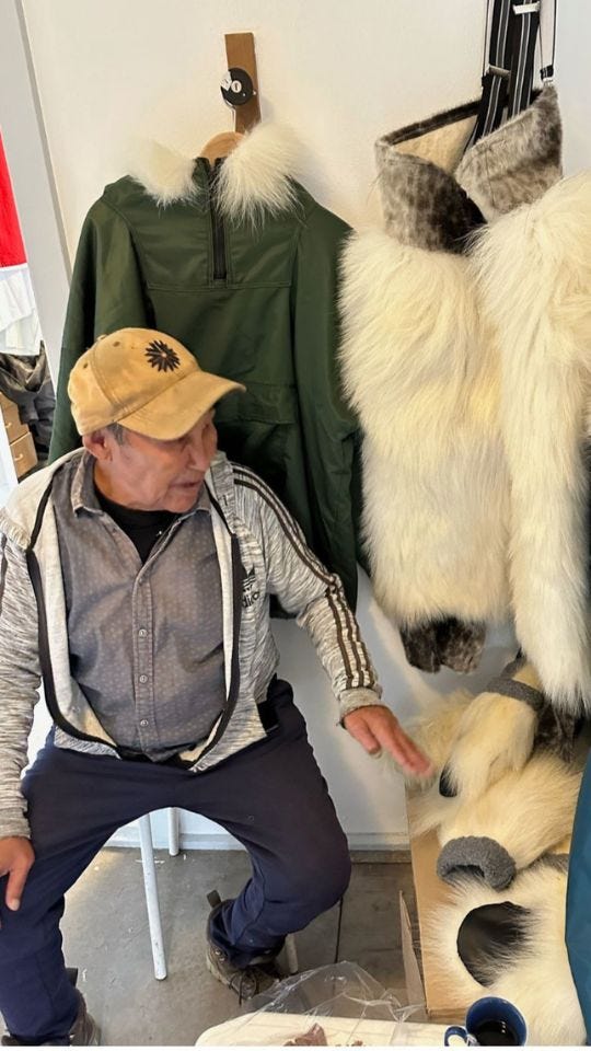 An older man wearing a tan cap and a gray track jacket sits in a small shop. Behind him and to his side hang polar bear boots and mittens with white fur, plus a green pullover with a fur-trimmed hood. Various straps and hooks on the wall.