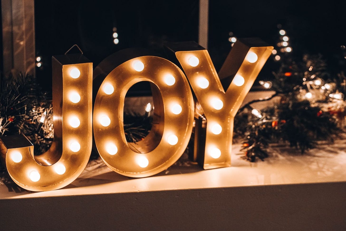 A metal-framed light display spelling JOY is sitting on a counter with various Christmas-themed greenery. A metal-framed light display spelling JOY is sitting on a counter with various Christmas-themed greenery.