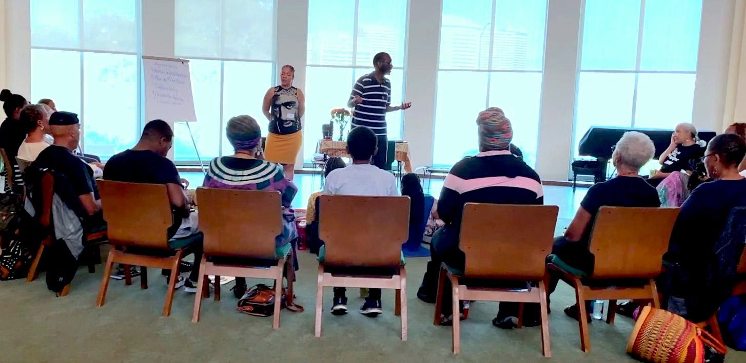 Kamal and co-facilitator stand before a circle of participants at a Healing Black Lives retreat. Behind them is a row of floor to ceiling windows.