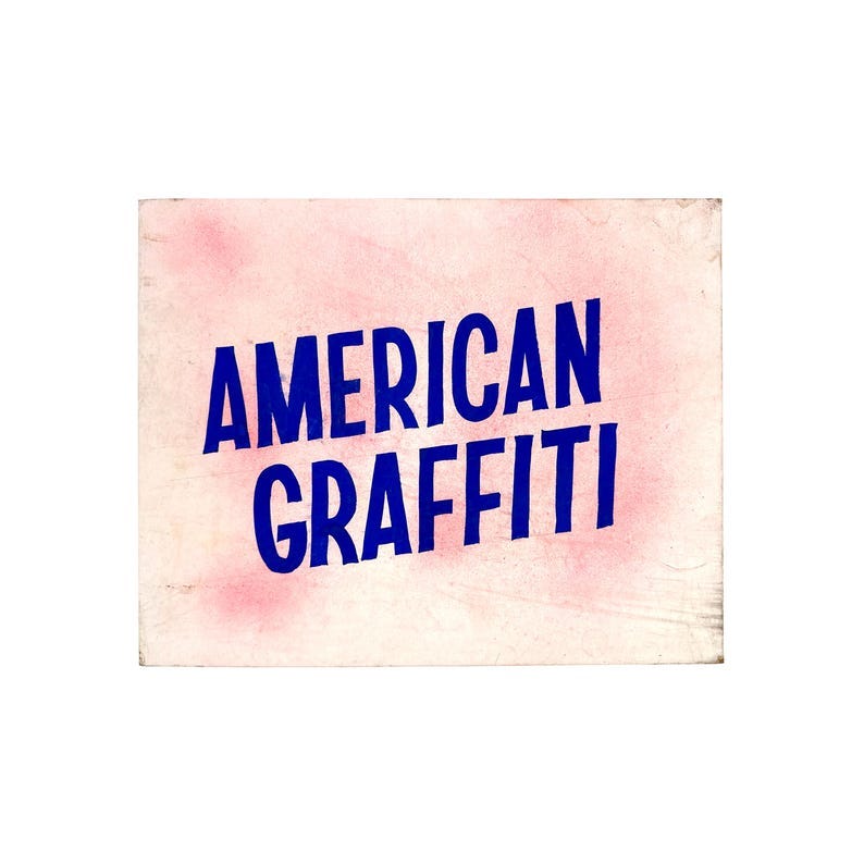 May include: A rectangular sign with the words 'AMERICAN GRAFFITI' in large, bold, dark blue letters. The text is diagonally oriented across the sign. The background is a light pink color with darker pink blotches. The sign has a slightly aged appearance.