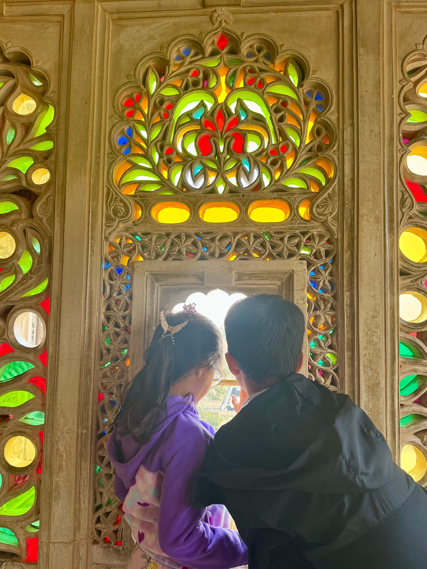 Two children looking out through an ornate stone lattice window with multicolored glass.