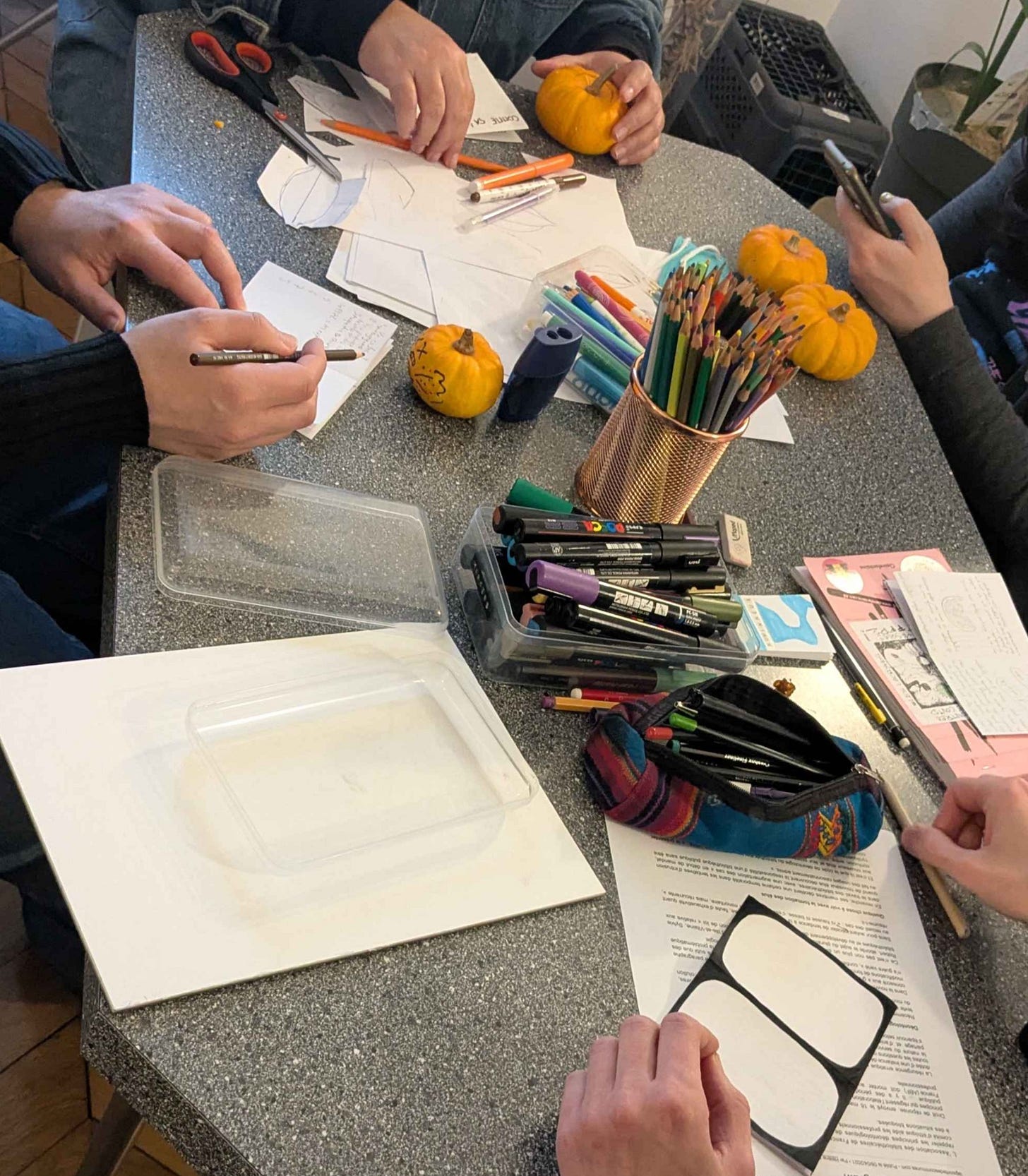 Photo d'une table où se trouvent ds feuilles, des crayons, des feutres et des petites citrouilles, entourées des mains des personnes qui participent à l'atelier