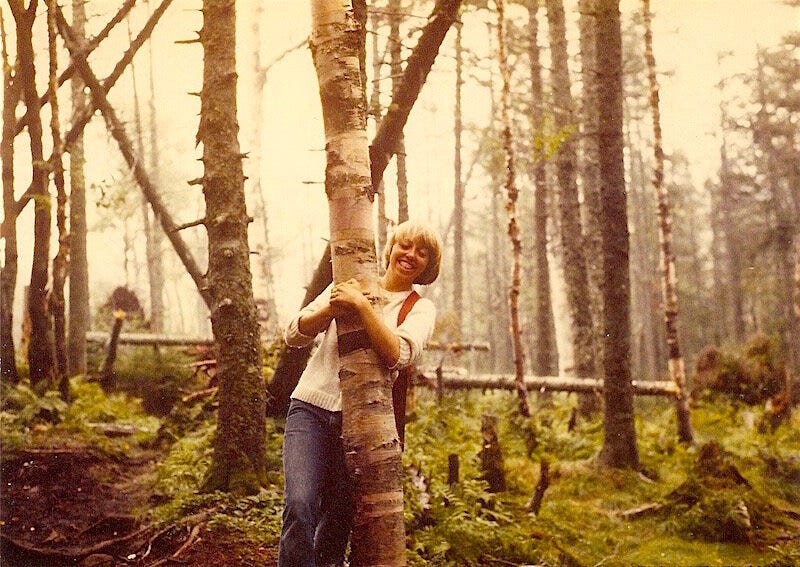 College-age girl in white sweater and jeans hugging a tree, smiling, in northern forest of moss and bare trees