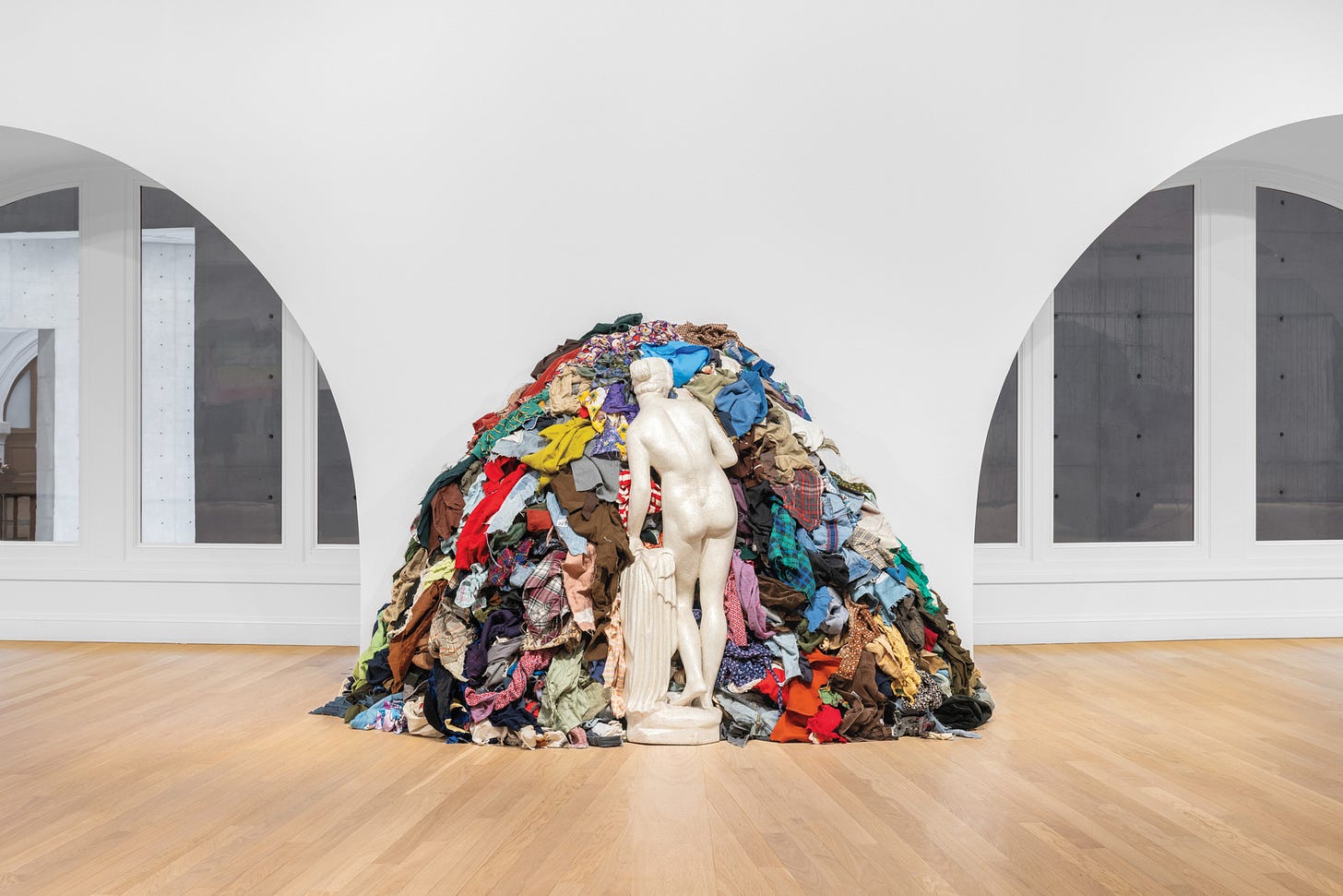 Sculpture of a nude woman made with a white material. She is standing in contraposto and facing a pile of used clothes.