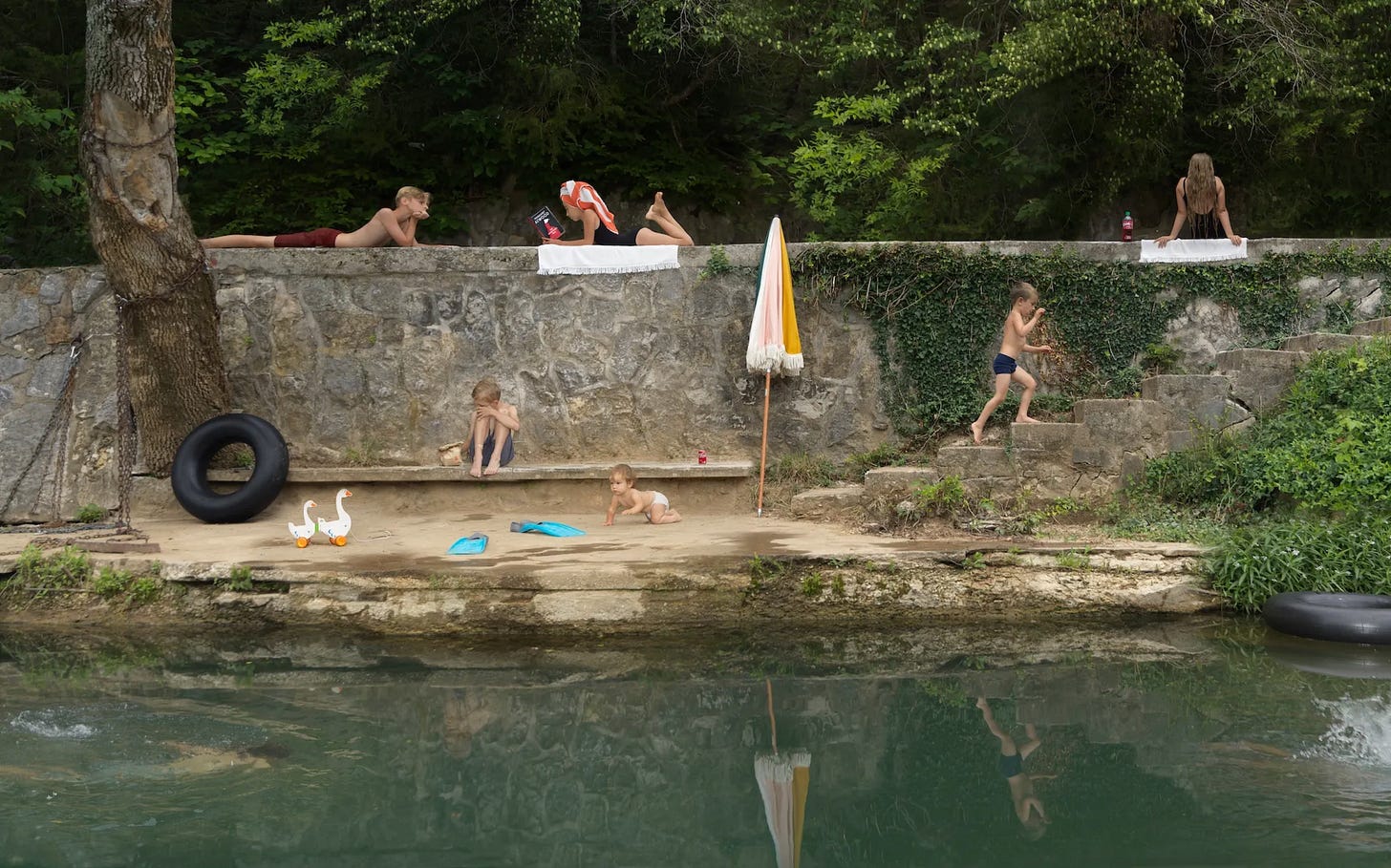"Children playing and relaxing by a stone wall near a river. Two children lie on top of the wall reading, while three children play on the sandy area below, with one child climbing steps and another sitting near the water. A baby is crawling on the sand. A black tire swing hangs from a tree, and a yellow and white umbrella is planted in the ground."