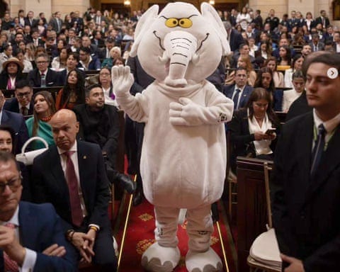 A person in an elephant mascot costume waves while standing in an aisle of a formal legislative chamber A person in an elephant mascot costume waves while standing in an aisle of a formal legislative chamber