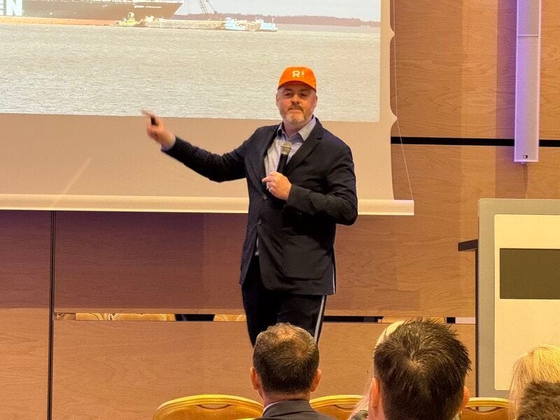 Christian standing on stage pointing to a screen while wearing an orange baseball cap, in front of an audience.