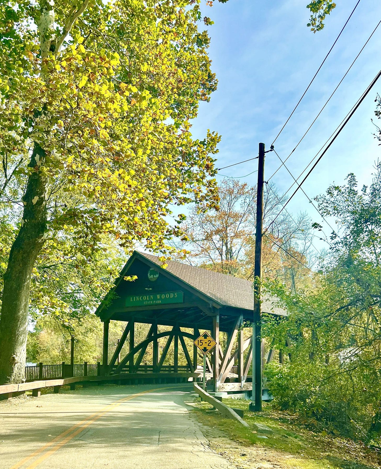 Entrance to Lincoln Woods State Park in Rhode Island, featuring a rustic wooden covered bridge framed by green autumn trees, with sunlight casting shadows on the road and a height clearance sign in the foreground — a scenic gateway to nature and forest trails.
