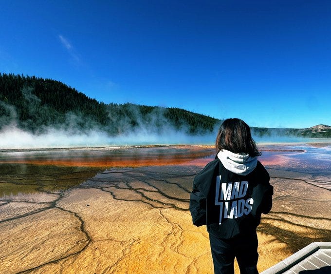 A person wearing a black hoodie with "MadLads" text on the back stands on a boardwalk. The scene shows a colorful thermal pool with orange and yellow hues, surrounded by cracked, dry ground. Steam rises from the water, and forested hills are visible under a clear blue sky.