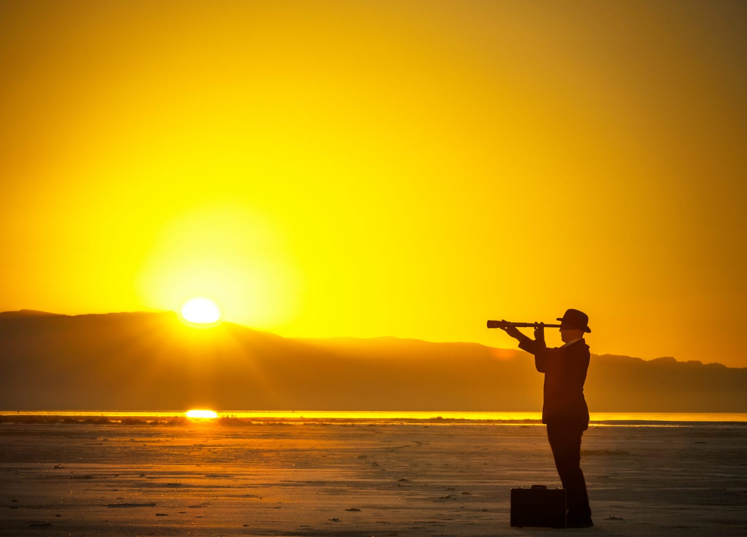A man in a suit and hat stands on the beach at sunset, looking through a telescope toward the horizon, with the sun glowing behind distant mountains.