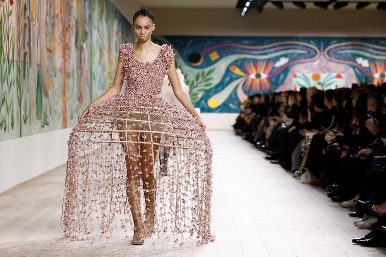 Uma modelo está desfilando em uma passarela, usando um vestido de design inovador, que é amplo e possui uma estrutura de rede. O fundo apresenta uma parede decorada com murais coloridos, enquanto uma plateia observa atentamente.
Uma modelo está desfilando em uma passarela, usando um vestido de design inovador, que é amplo e possui uma estrutura de rede. O fundo apresenta uma parede decorada com murais coloridos, enquanto uma plateia observa atentamente.
