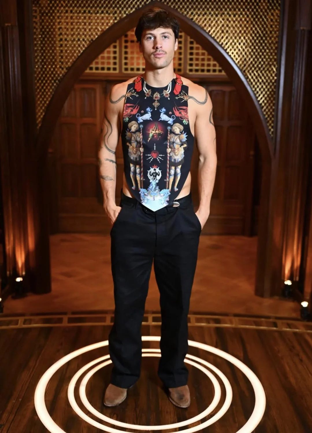 Rob in an ornate printed vest with religious imagery over a bare chest, belted shoulder straps, black trousers, and brown leather boots.