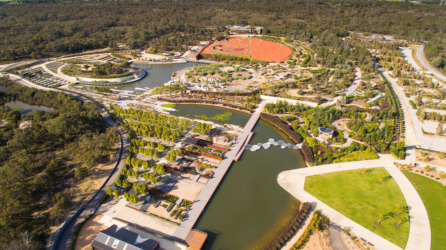 Aerial view of Cranbourne Botanics. Lakes, small gardens, pathways and a large patterned area of red earth are surrounded by forest.