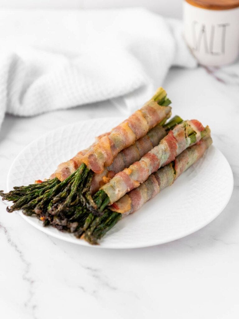 Bacon-wrapped asparagus spears arranged on a white plate atop a marble surface.