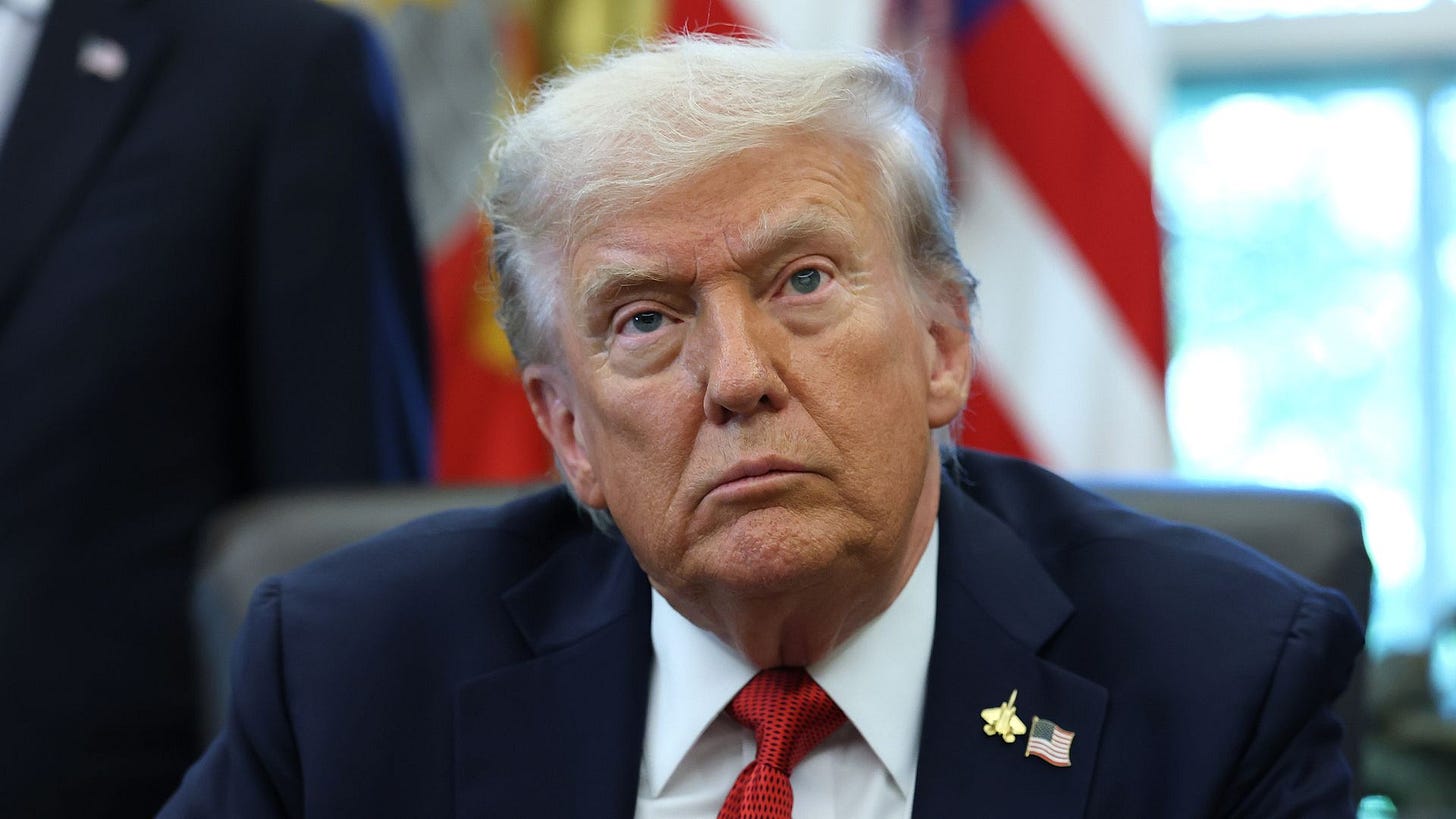 President Trump, wearing a navy jacket with a US flag pin, and a gold plane pin white shirt and red tie, stares into the distance while sitting on a black chair  in the Oval Office at the White House.