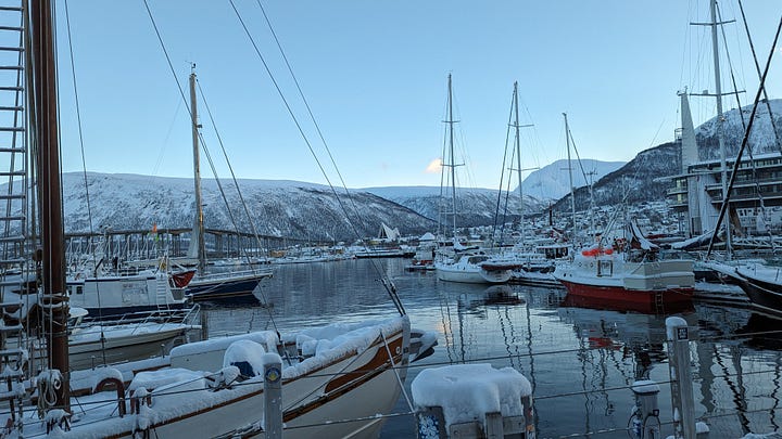 A woman stands with her arms upstretched, delighted, atop Fjellheisen mountain in the snow. Also, Tromso harbour, boats clinking on the still, glassy water of the fjord.