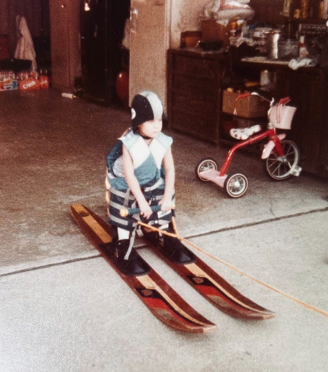 Young child stands in the driveway on wooden water skis, wearing an oversized life jacket and helmet, gripping a practice rope, capturing a playful moment of determination and admiration for his uncle’s water skiing legacy and the joy of family mentorship.