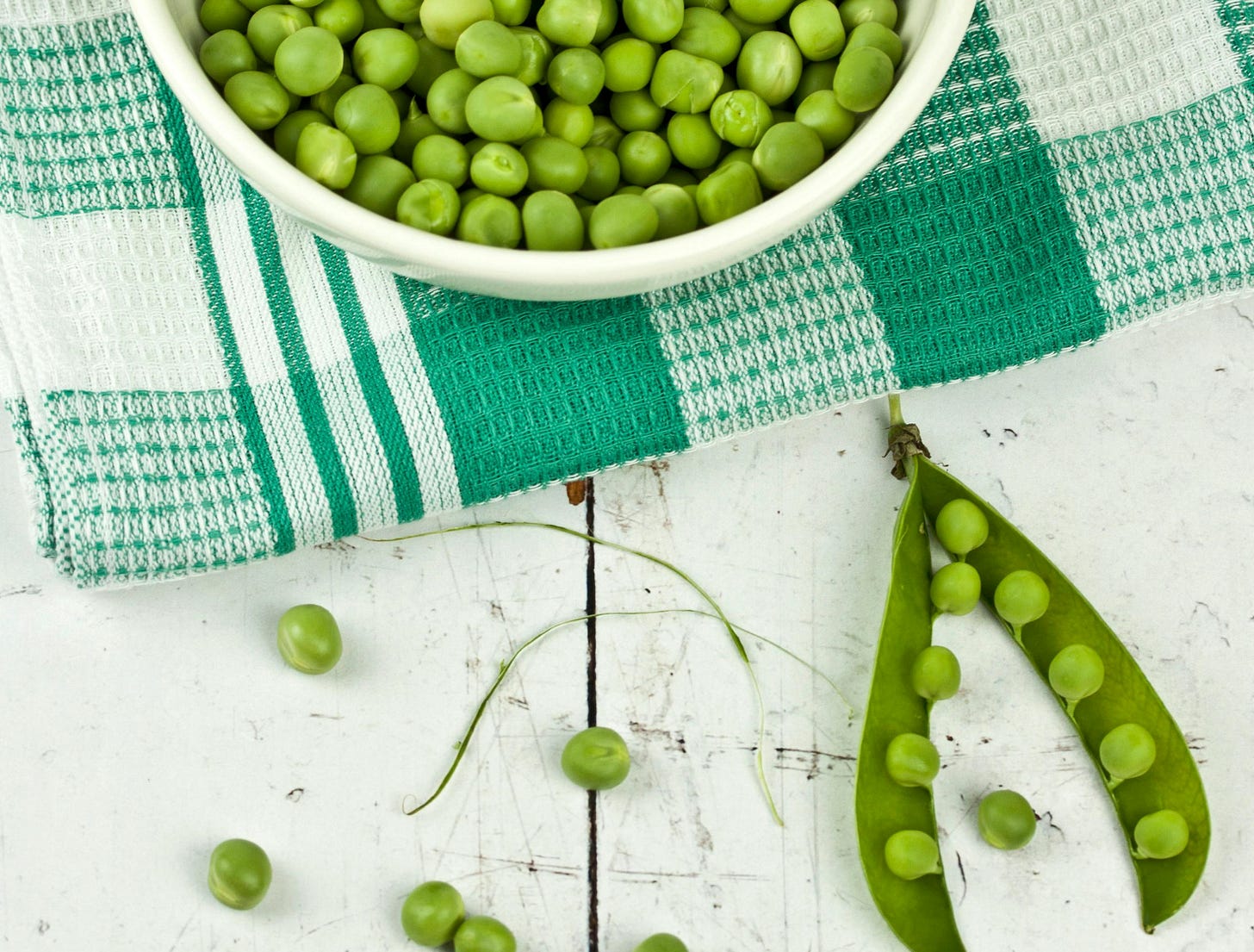 Bowl of fresh peas on folded towel, with open pod full of peas on the table